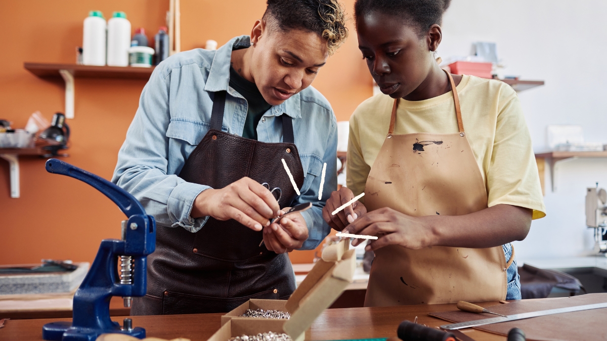 Une porteuse de savoir-faire montre un geste de maroquinerie à une visiteuse dans son lieu de production